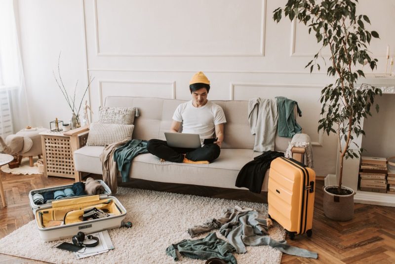 Man in a yellow beanie working on a laptop with open suitcases in a cozy living room.
